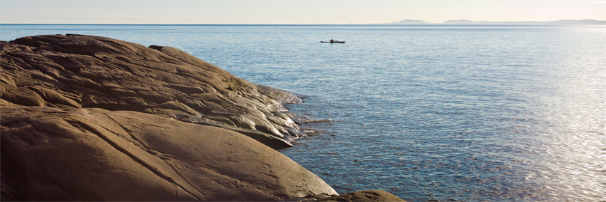 Prise de vue terrestre sur des rochers montrant une mer calme où navigue un kayak au loin.