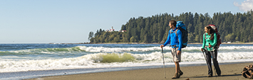 Two hikers walking along the beach with lighthouse in background on the West Coast Trail.