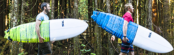 Deux jeunes hommes transportant des planches de surf le long d’une route avec la forêt pluviale en arrière-plan.