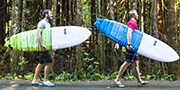 Deux jeunes hommes transportant des planches de surf le long d’une route avec la forêt pluviale en arrière-plan.