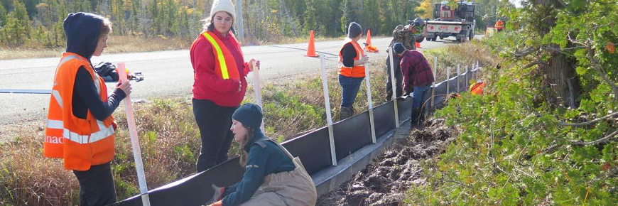 People in a roadside ditch adjusting a low barrier secured with stakes.