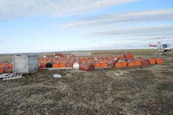 Fuel barrels collected on the tundra with a helicopter nearby.