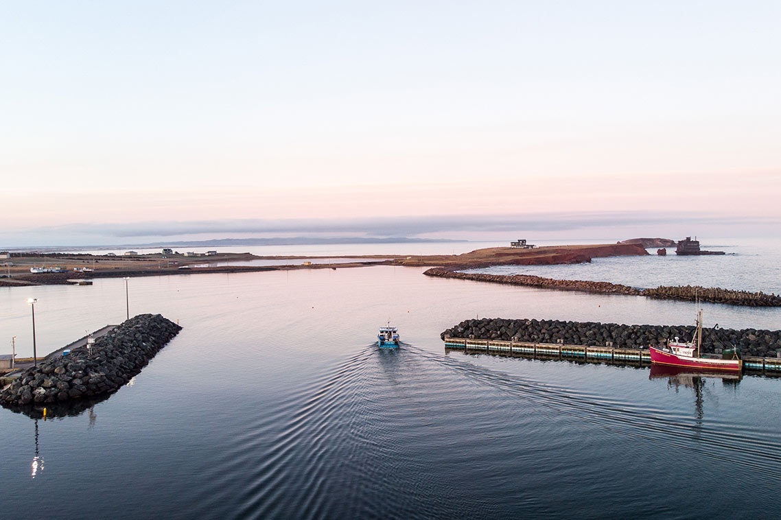 A fishing vessel moves slowly through the flat water as it exits a port area. Different barriers, breakwaters, and another red and white boat are visisble in the mid-ground and the Magdalen Islands' coastline sits in the far-ground.