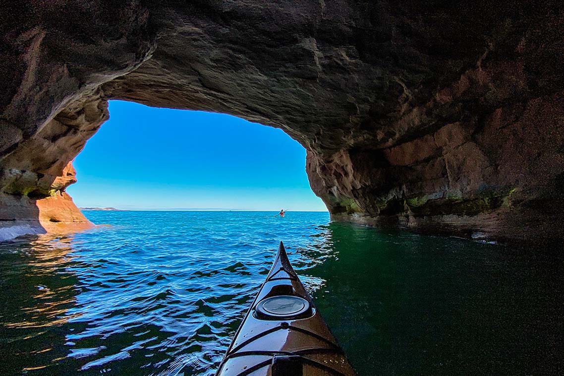 The bow of a kayak pushes forward in Gult of St-Lawrence waters. Coastal rocks stretch overhead and in front, except for a rounded opening revealing a second kayaker and more ocean ahead.
