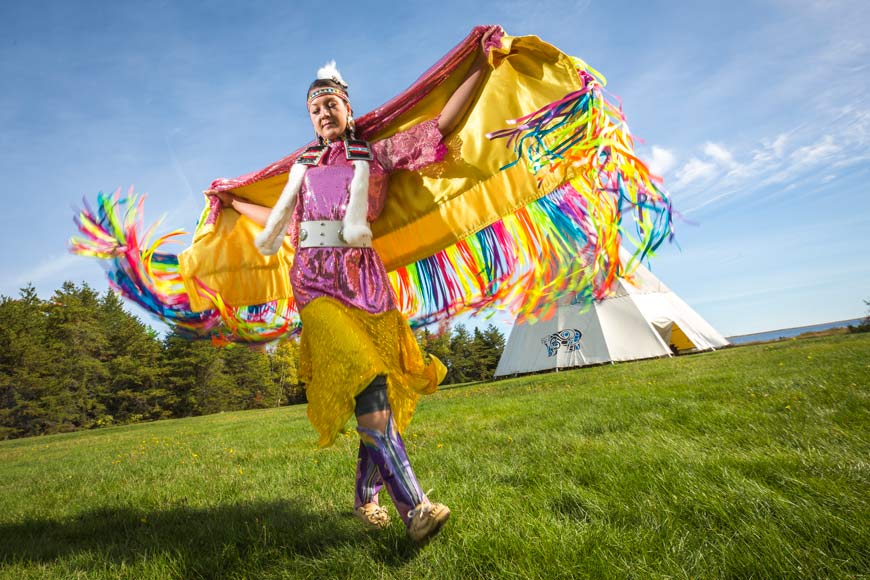 Indigenous woman wearing fancy shawl, dancing in the green grass in front of large Wigwam.