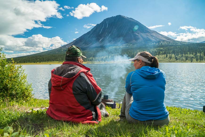 A man and a women sit in front of a lake while steam raises in front of them from a pot with a large mountain directly across.