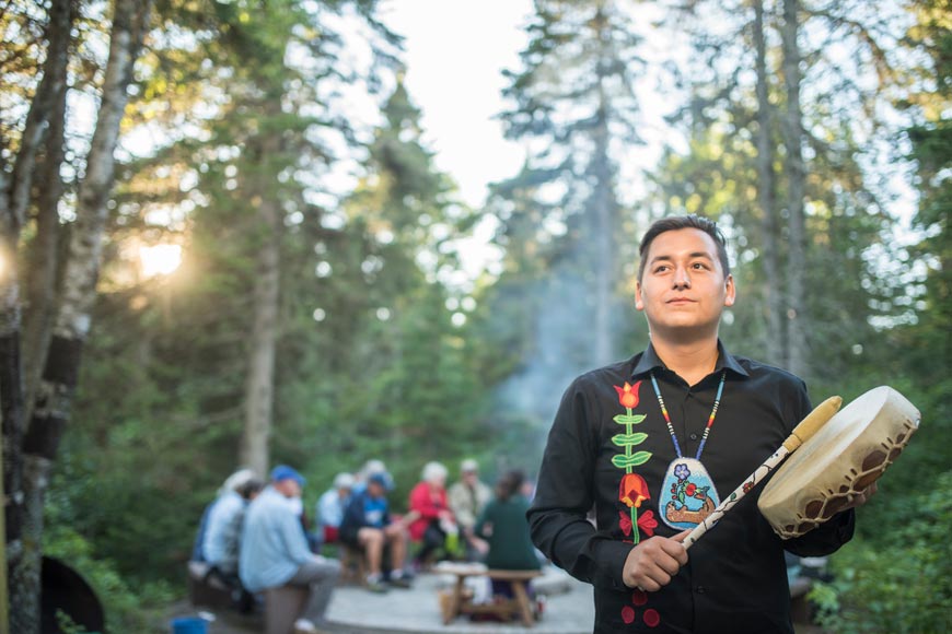 Homme indigène vêtu en linge noir avec un collier en couleur joue son tambour traditionnel devant un feu.