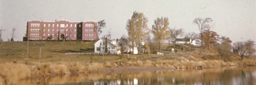 Shubenacadie Residential School buildings and some trees reflected in the Shubenacadie River on a clear day.