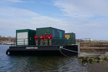 Four people in red outerwear stand on a flat barge in front of green metal storage units