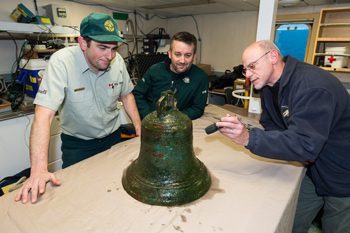 Three men looking at a ship’s bell on a table.