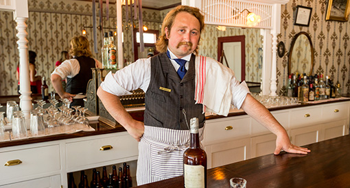 Un guide en tenue typique de barman de l'époque de la ruée vers l'or, servant un verre au saloon Red Feather, au lieu historique national du Complexe-Historique-de-Dawson