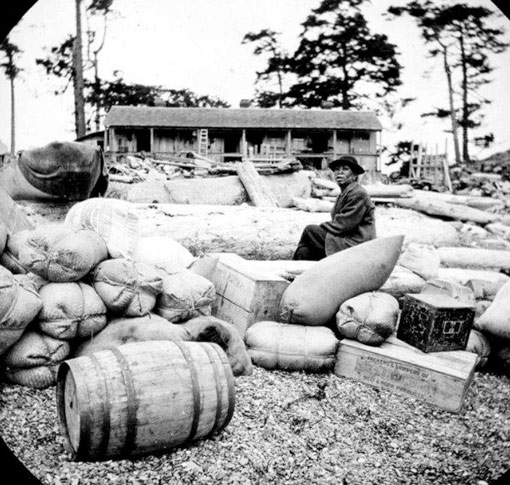 Black and white photo of men on a beach with barrels, boxes and other supplies being delivered.