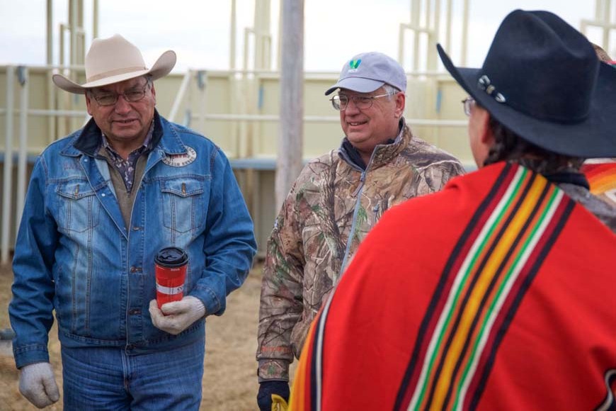 Three men are gathered in a bison compound. One holds a coffee, one is smiling, one has his head turned away.