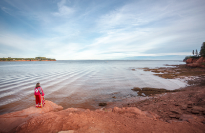 Une femme portant une longue robe rouge, tenant un tambour à main, se tient sur le rivage tout en regardant dans l’eau.