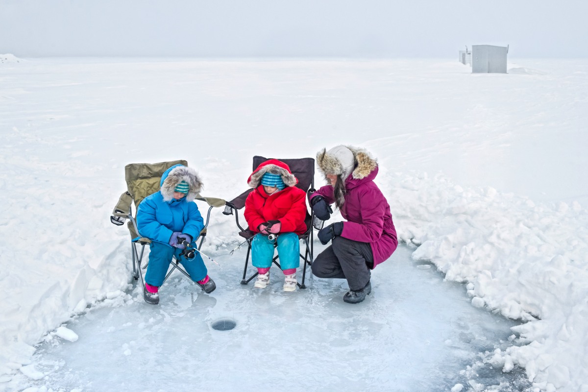 Une mère et ses deux enfants sont emmitouflés sur un paysage plat et recouvert de neige, alors qu’ils sont réunis autour d’un trou de pêche sur glace.