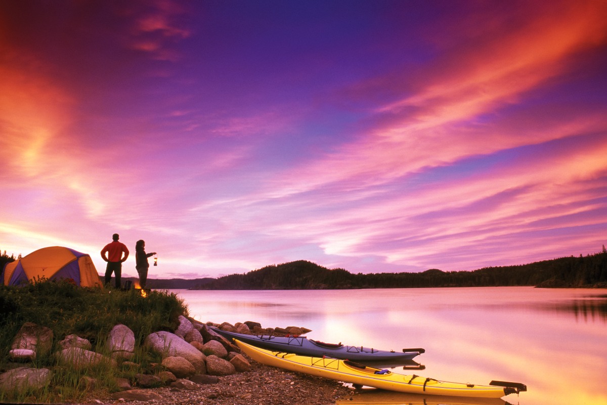 Deux personnes debout près de leur tente et de leurs kayaks regardent les eaux calmes au coucher du soleil.