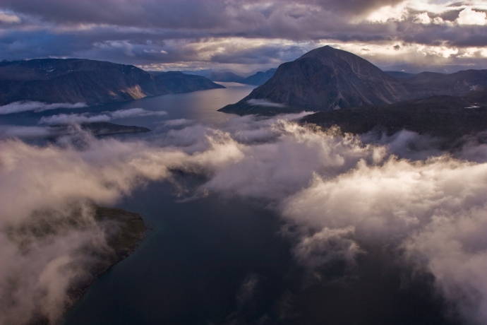 Vue aérienne, au-dessus de nuages bas, d’un fjord et d’un paysage montagneux.