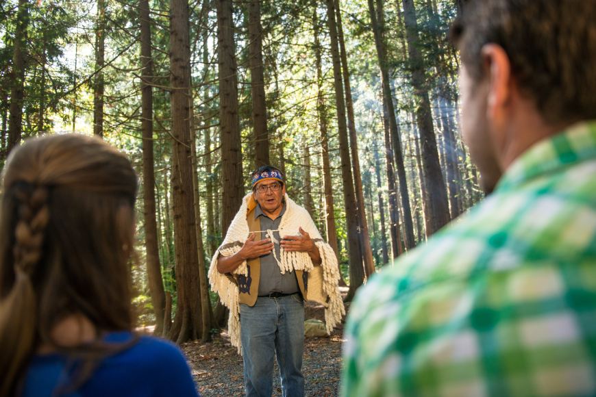 A person standing in the woods engages two people through storytelling.