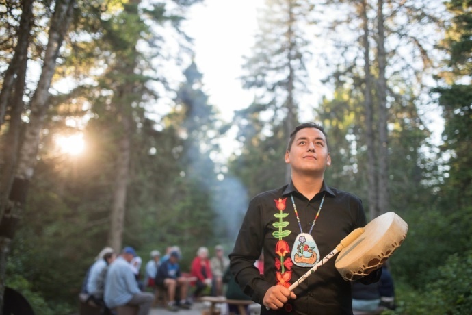 A person holding a hand-drum looks up while a group of people sit around a fire in the background.