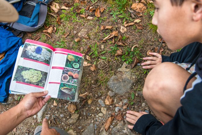 A close up of a child kneeling on the ground, as they look at a guidebook held by another.