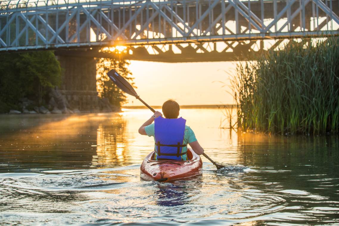 A person wearing a lifejacket sits on a kayak while paddling toward marsh grasses and a bridge overhead.