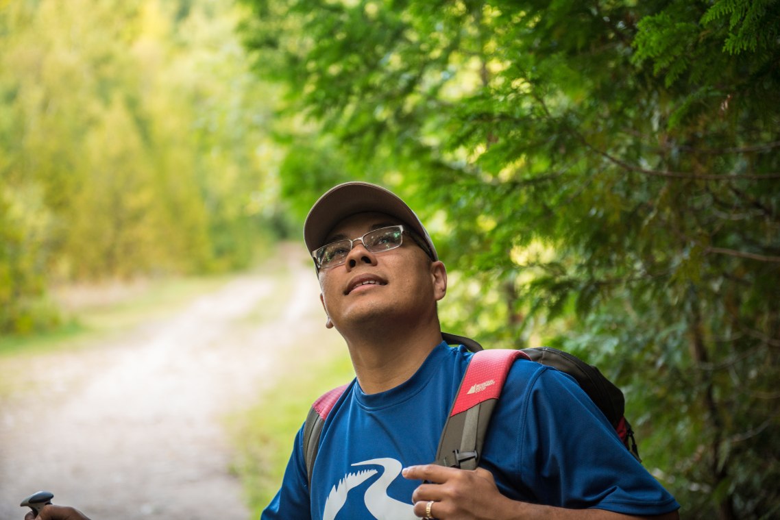 A close-up of a person smiling stands while looking upward on a forested trail.
