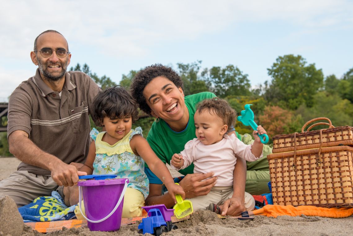 A family with two young children sit smiling on a picnic blanket while the children play in the sand.