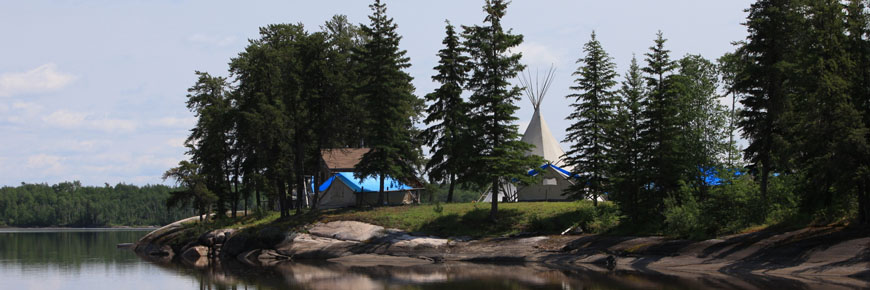 Tent structures, including a teepee, are erected among evergreen trees at the side of a lake.