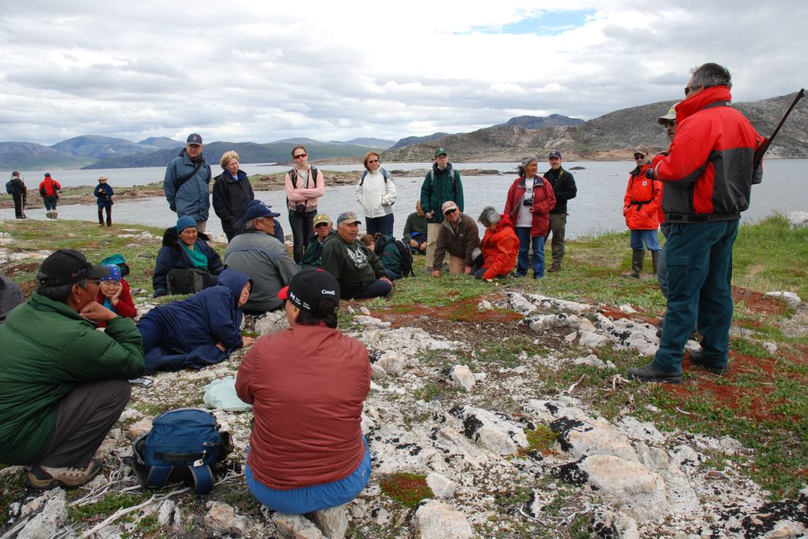 A group of people sit and stand on grass near a waterbody surrounded by mountains as they listen to another talking to the group.