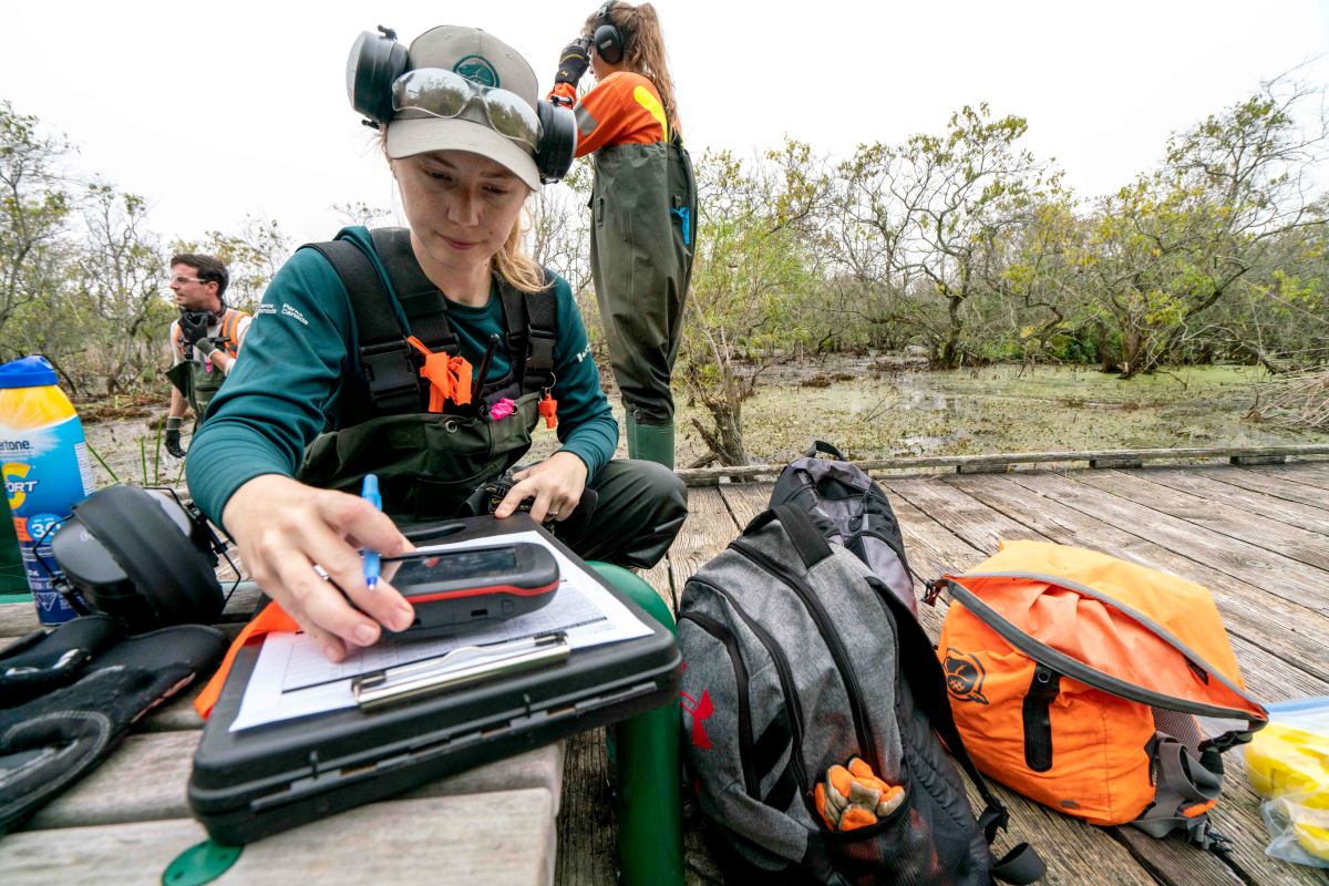 A Parks Canada staff member checks a GPS device and paperwork while other staff work in the background.