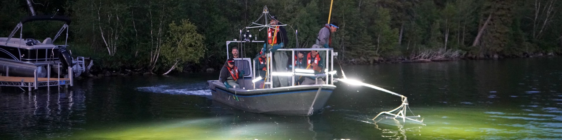 Crew aboard an electrofishing boat at dusk illuminate the lake as they trail suspended gear through the water.
