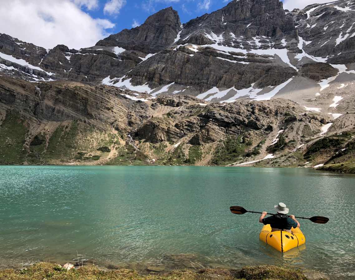 A person sits in a yellow inflatable boat on a lake surrounded by snowcapped mountains and hillsides.