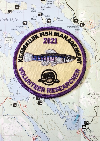A fabric badge with the words “Kejimkujik fish management, 2021, volunteer researcher” along with an image of a fish and the Parks Canada logo is shown on top of a map.