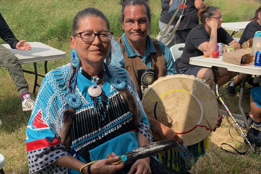 Une femme autochtone en tenue de cérémonie est assise à côté de son partenaire autochtone qui tient un tambour à main.