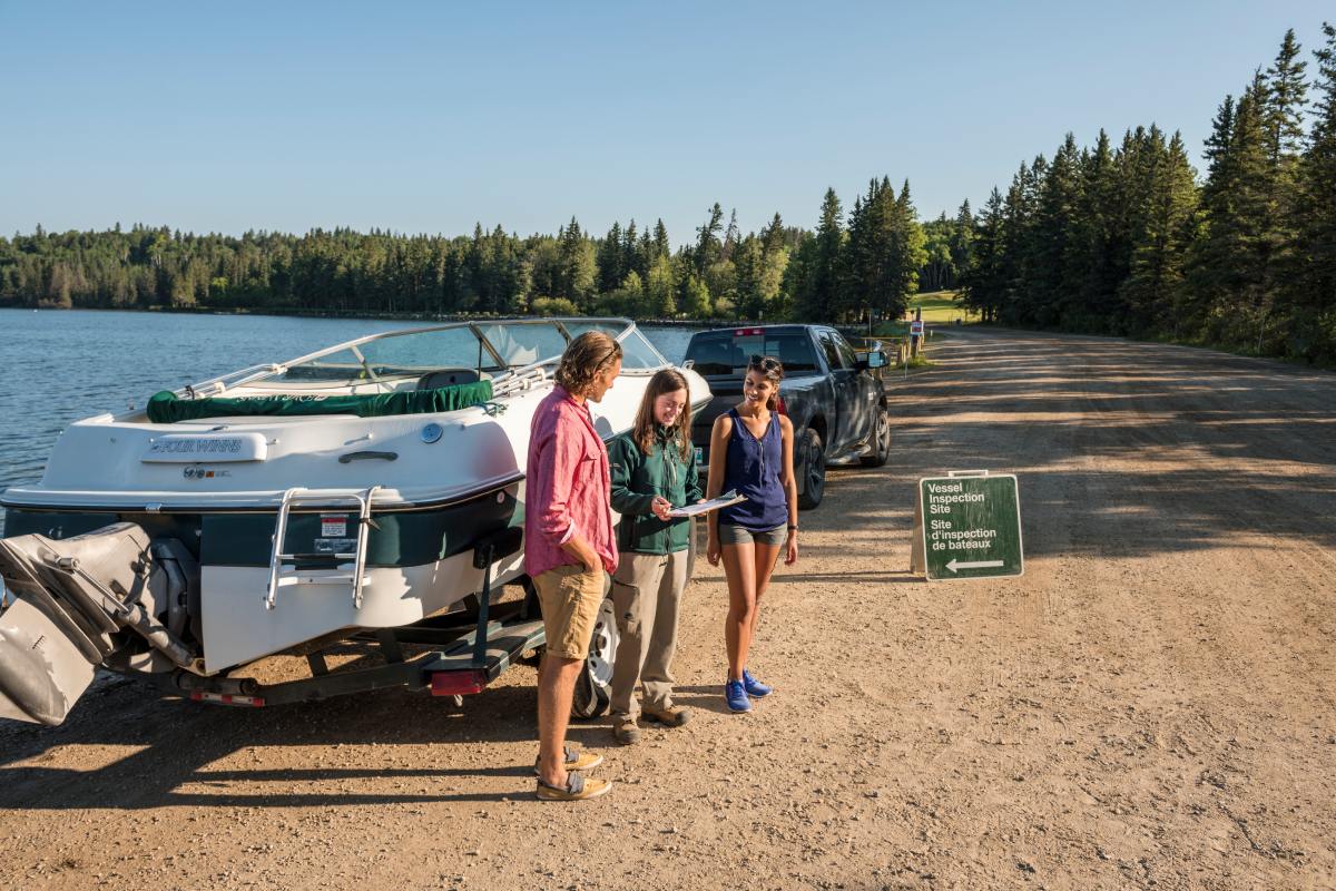 Un membre du personnel de Parcs Canada passe en revue une liste de contrôle avec deux visiteurs et leur bateau à un poste d’inspection au bord du lac.