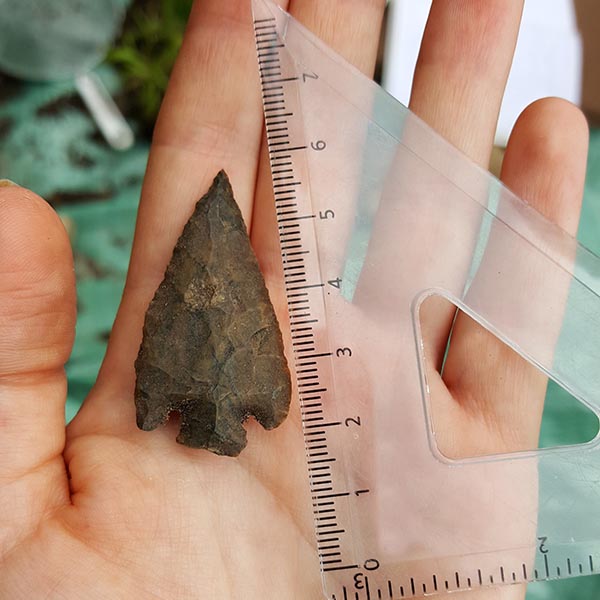 A stone arrowhead is held in the palm of a hand. A measuring tool sits next to the arrowhead indicating a length of about 5 centimeters.