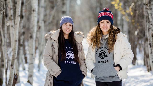 Deux femmes se tiennent debout dans la forêt enneigée et portent des cotons ouatés et des tuques de Parcs Canada