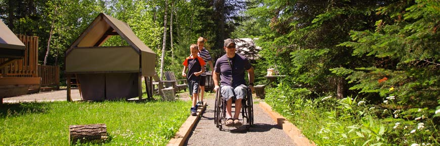 Un père en fauteuil roulant avec ses deux enfants sur le sentier accessible à côté d’une tente oTENTik dotée d’une rampe d’accès.