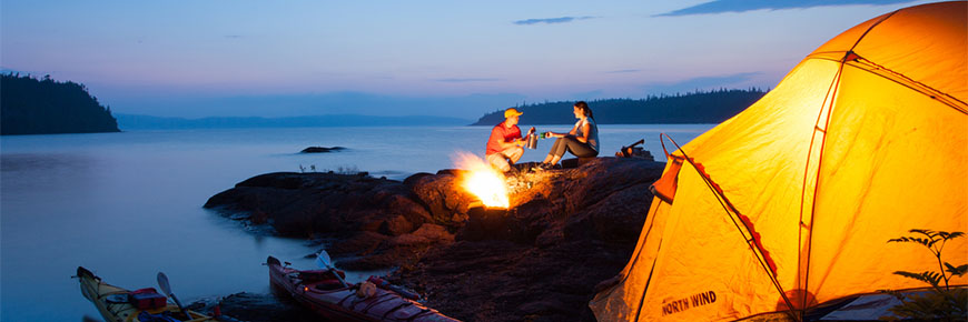 A couple relaxes by the fire on Harry Island next to the water.
