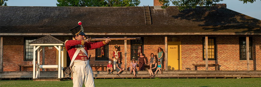 Visitors sit outside on the porch of one of the historic buildings of Fort Malden while a costumed interpreter fires a musket