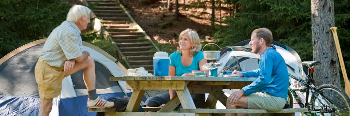 Trois adultes profitent d’un repas sur une table de pique-nique au terrain de camping Point Wolfe.