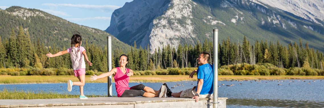 A young girl runs to her parents on a dock with Mount Rundle in the background.