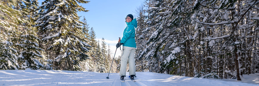 Une femme fait du ski de fond sur un sentier dans une forêt de conifères.