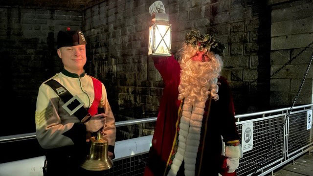 Father Christmas and a Highlander holding a lantern in front of the Halifax Citadel entrance.
