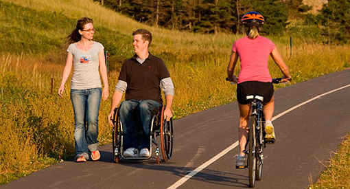 A man in a wheelchair and a woman walking on the Gulf Shore Way East Trail. A cyclist travels in the opposite direction.