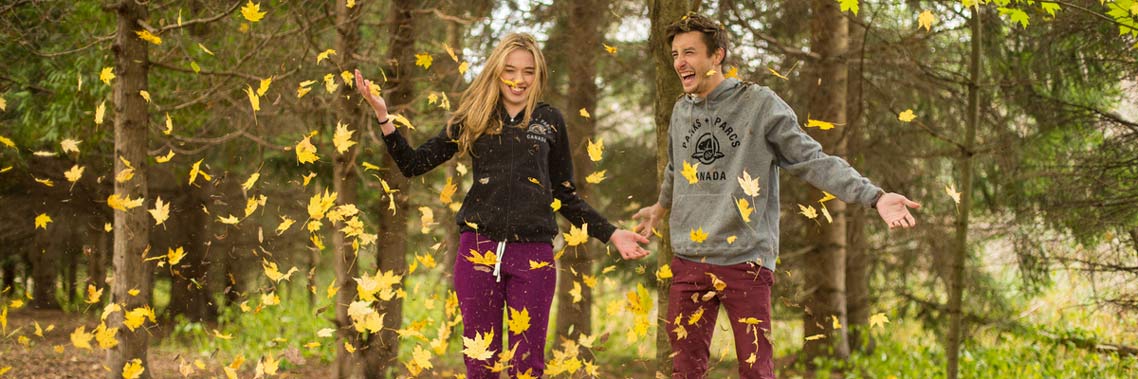 A couple laughs as leaves fall around them in the forest.