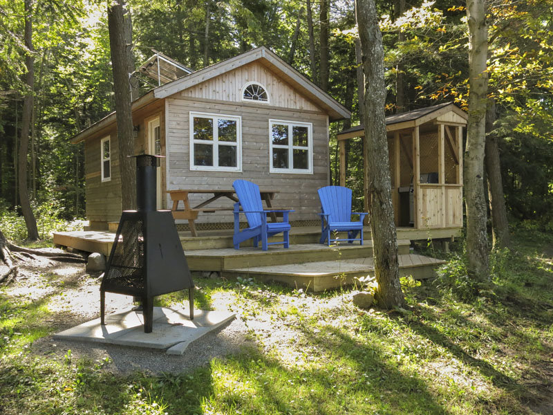 Des chaises bleues et un barbecue sont installés sur la terrasse d'un chalet rustique. Un foyer extérieur est à proximité.
