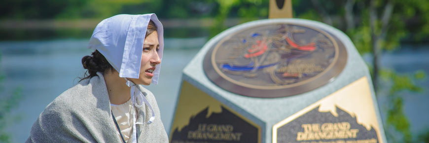 Une femme en costume d’époque devant un monument sur le Grand Dérangement.