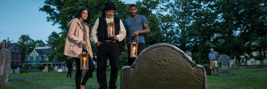 A young couple holding lanterns stands with a costumed guide at Garrison Cemetery.