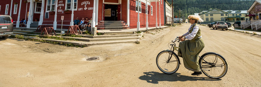 Une femme en costume d’époque fait du vélo devant un bâtiment historique de Dawson City.